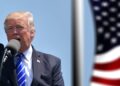 Donald Trump hablando con un microfono junto a una bandera de Estados Unidos