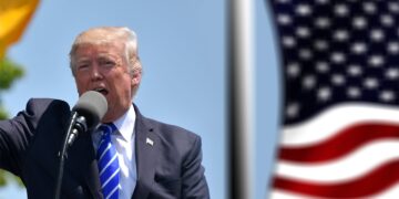 Donald Trump hablando con un microfono junto a una bandera de Estados Unidos
